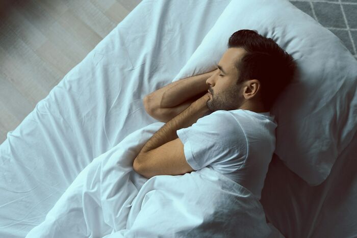 Man lying in bed with eyes closed under white sheets, symbolizing feelings related to weaponized incompetence.