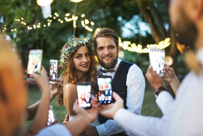 Bride and groom smiling as guests capture photos with smartphones, illustrating tacky wedding stories with modern distractions.