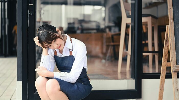 A young barista in an apron looks distressed, sitting outside a cafe. Rage quit or feeling overwhelmed at work.