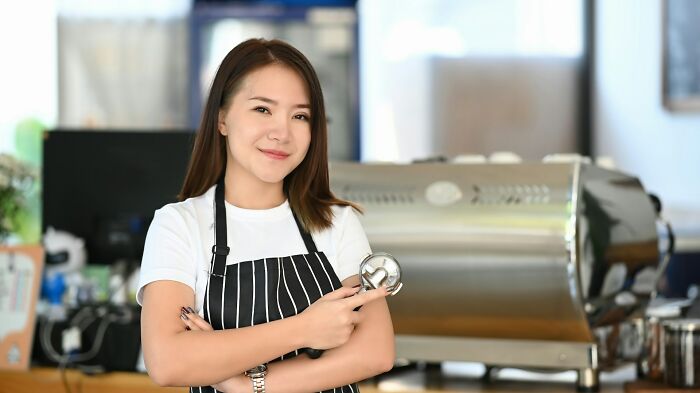 Young woman in striped apron holding a coffee scoop, smiling confidently in a bright café, illustrating karma satisfaction.