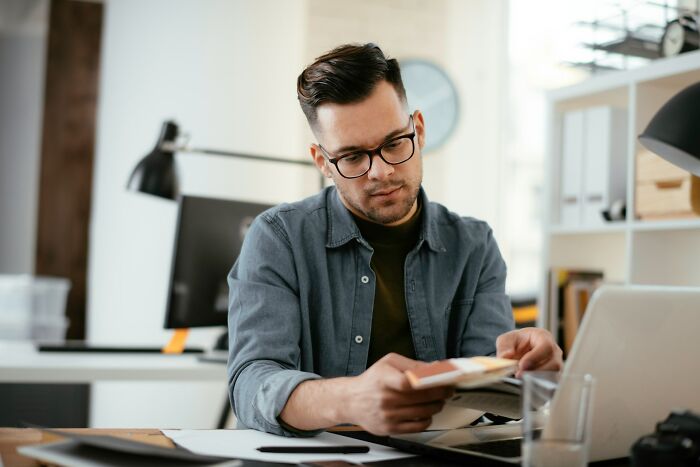 Man wearing glasses and a denim shirt reading a book at his desk, illustrating stories about karma in various forms.