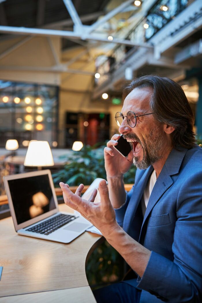 Man in glasses and blue suit yelling into phone while working, perhaps ready to rage quit his job.
