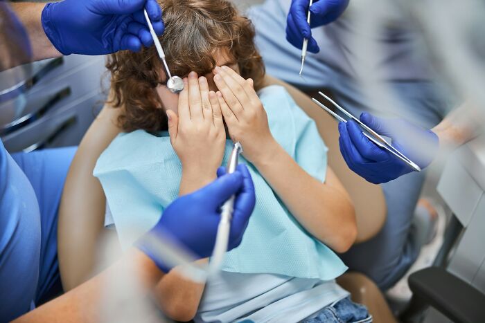 Child covering face nervously at a dental checkup surrounded by gloved hands with dental tools, parenting tips concept.