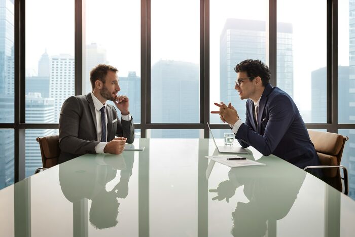 Two men in suits, one listening intently while the other gestures, discussing a rage quit in a modern office with city views.