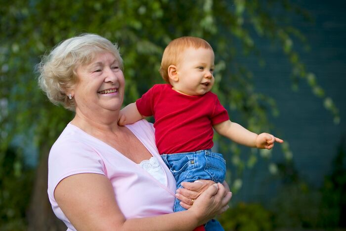 A smiling grandmother holds a young child in a red shirt who is pointing. Is your MIL the worst?