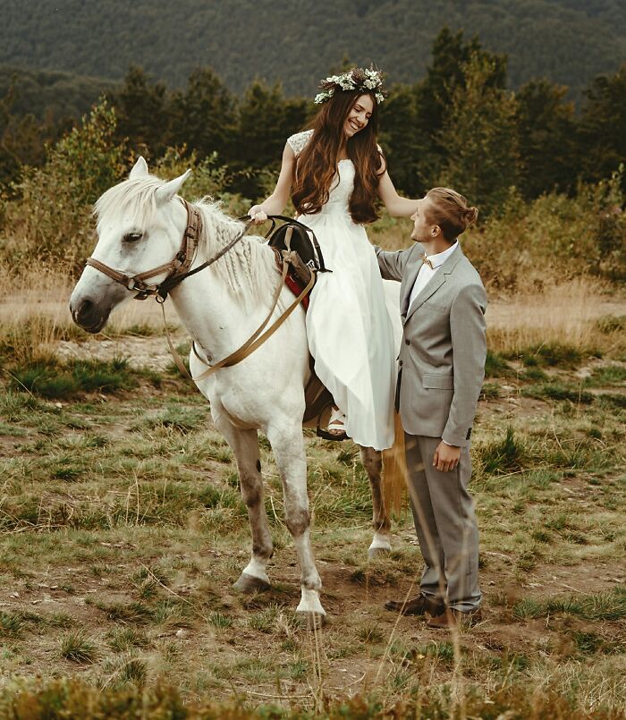 Bride in a white dress sitting on a white horse while groom in a suit stands beside her outdoors at a wedding.