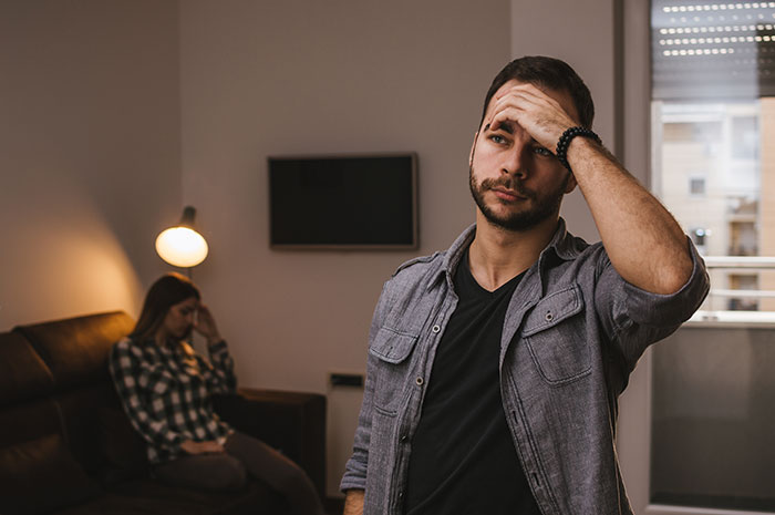 Man feeling like a science experiment at home, frustrated while woman sits in the background keeping a log of his words.