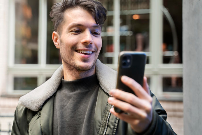 A smiling man in a green jacket with a fur collar looks at his phone, possibly involved in an affair.
