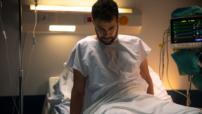 A man in a hospital gown, connected to an IV and oxygen, sits on a bed with a pained expression. Exposure reveals disturbing details.