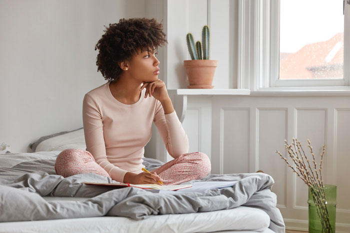 Woman thinking pensively on a bed, journal in lap. She considers unsettling details, possibly about an affair.