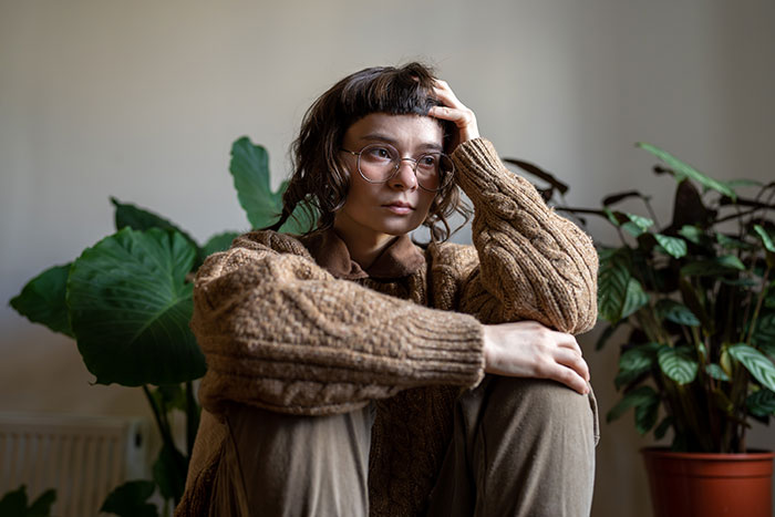 Woman sitting thoughtfully indoors, wearing glasses and a brown sweater, reflecting on relationship and feeling liberated.