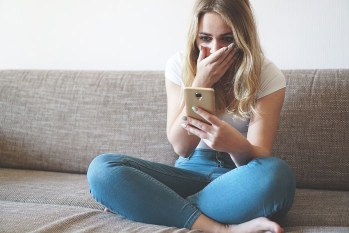 Woman shocked looking at phone sitting cross-legged on couch