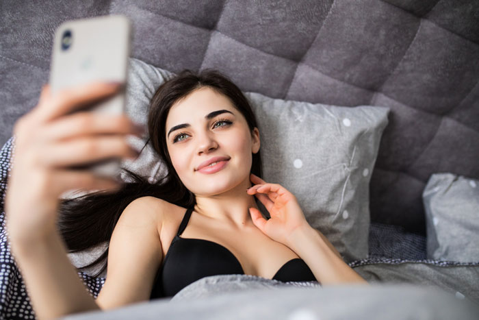 Young woman in black bra taking a selfie in bed, showing off piercings, highlighting social media and relationships.