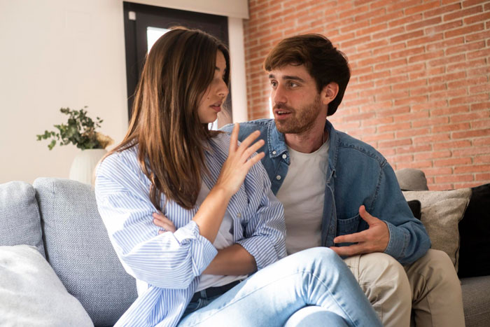 Couple having an intense conversation on a couch, woman showing off piercings as boyfriend reacts with concern.