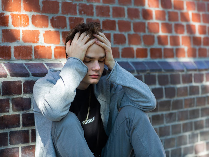 Young man sits against brick wall, holding his head in distress, reflecting upset boyfriend emotions from piercing and relationship issues.