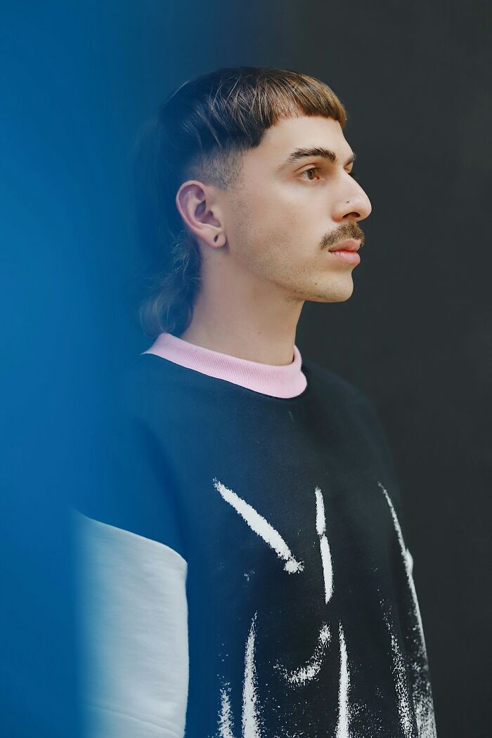 Young man with unique hairstyle and mustache wearing a black and pink sweater, showcasing intelligence and creative style.