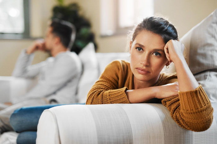 Woman looking upset on couch while man sits in background, illustrating a bro demanding sister&rsquo;s heirloom ring for proposal conflict.