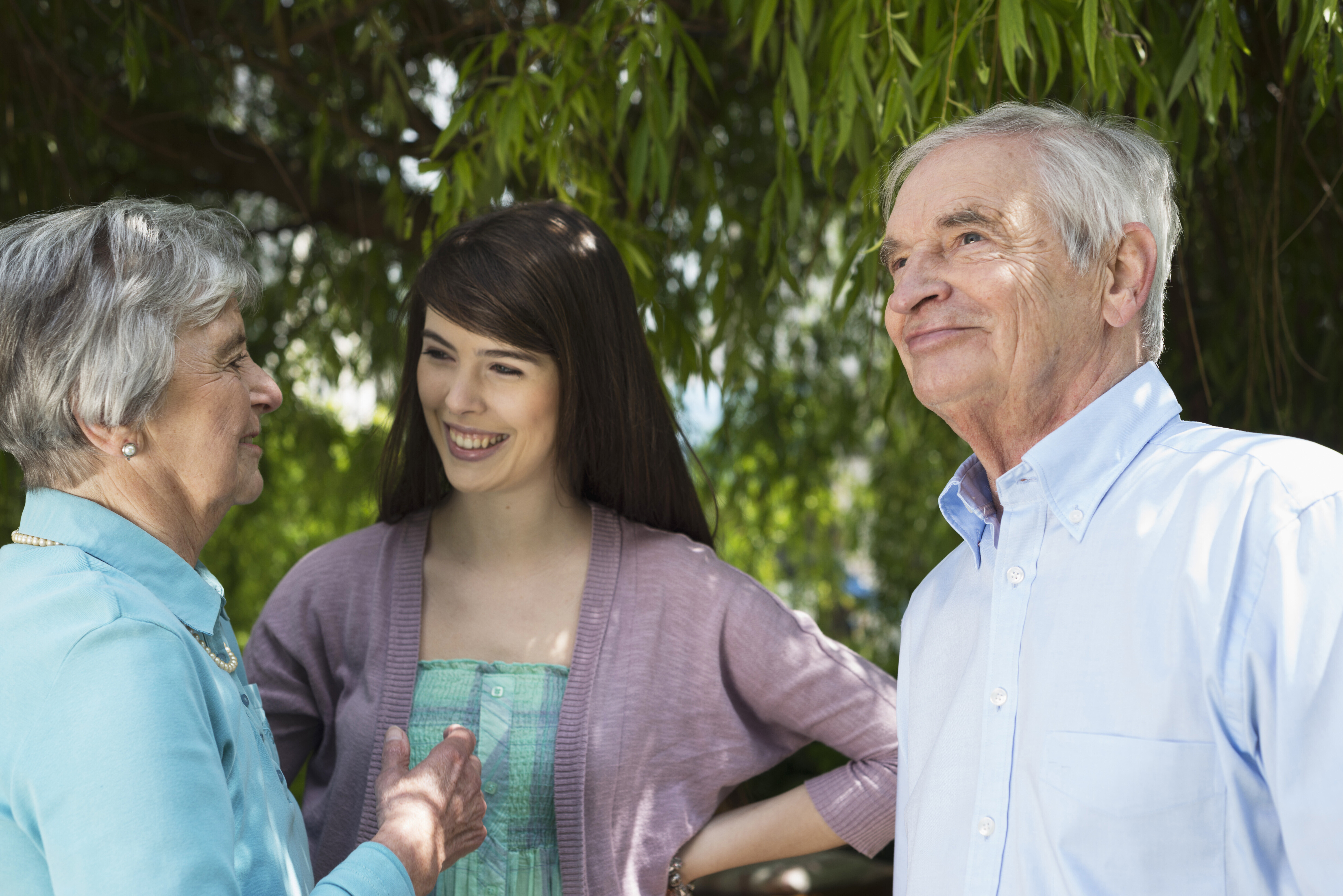Young woman smiling while talking with older couple outside under a tree, family and controlling friend discussion.