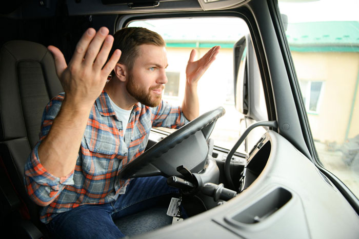 A frustrated driver in a truck, wearing a plaid shirt and beard, throws his hands up in anger, perhaps after being cut off. Driver thinks he&rsquo;s following guy.