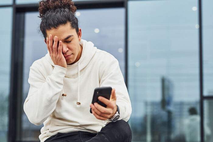 A man with a man bun, wearing a white hoodie, holds a phone and covers his face in distress, finding out he was being cheated on.