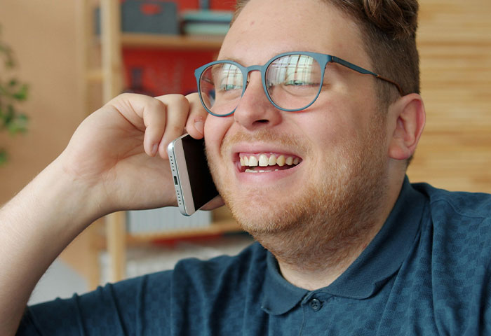 A man wearing glasses smiling and talking on a phone, representing harmless but effective revenge on jerks.