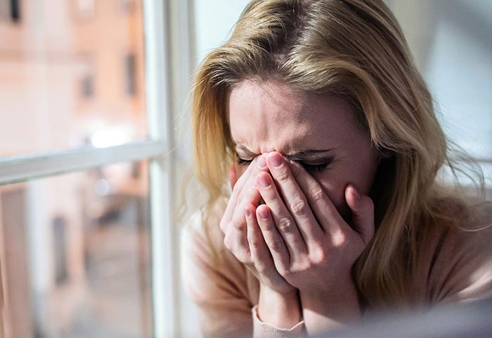 A distressed woman covering her face with hands near a window, symbolizing emotions in harmless revenge on jerks.