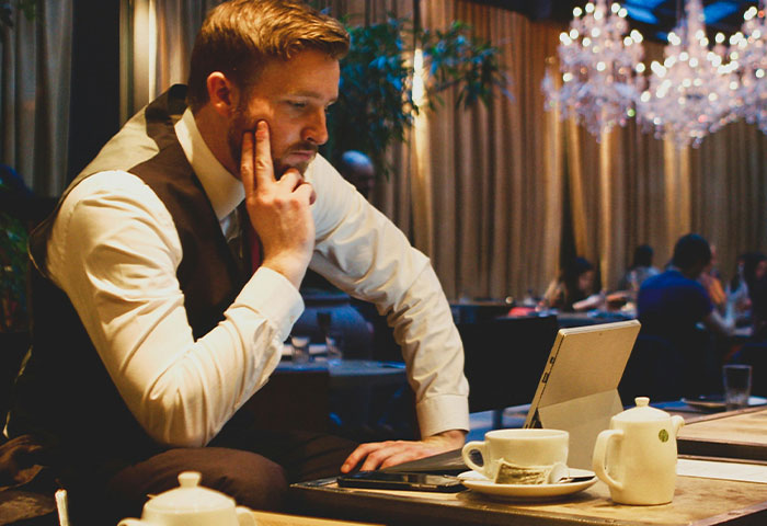 A man in a vest thoughtfully using a laptop in a cafe, illustrating harmless but effective ways to get revenge.