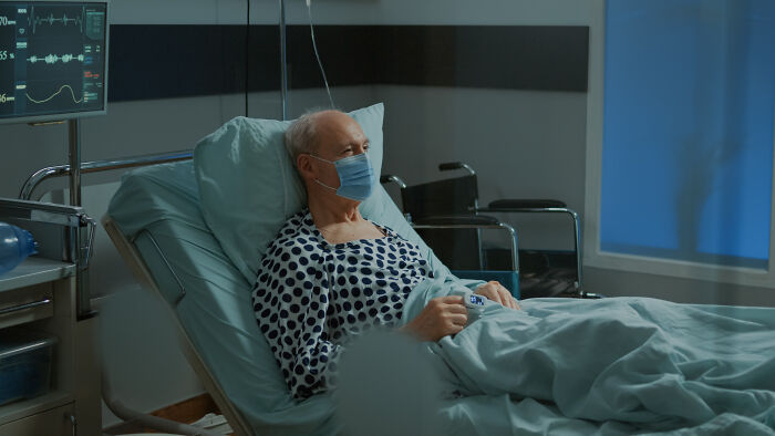Elderly patient wearing mask lying in hospital bed with heart monitor nearby, representing healthcare workers' care.