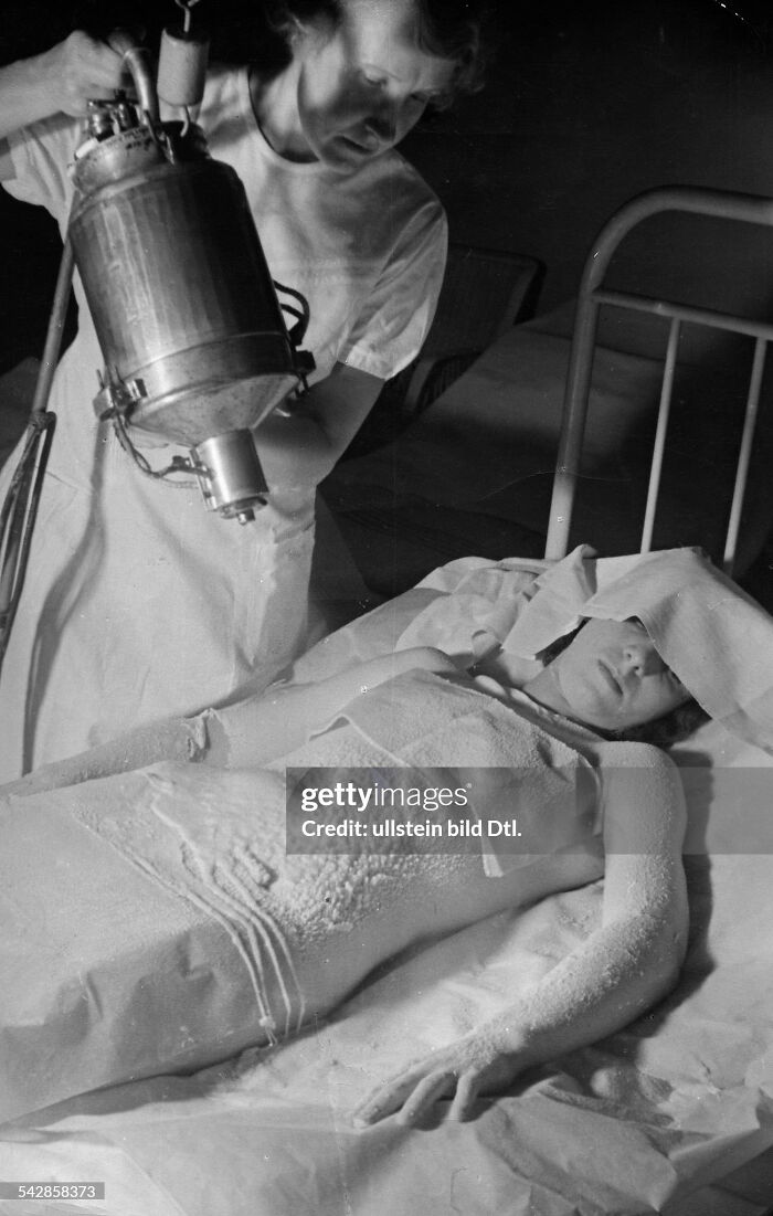 Nurse using vintage medical device to treat patient covered in medicinal substance in a historic healthcare setting.