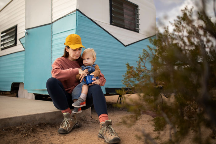 A woman in a yellow hat and toddler sitting outdoors by a teal and white camper, illustrating homeless ex and child.