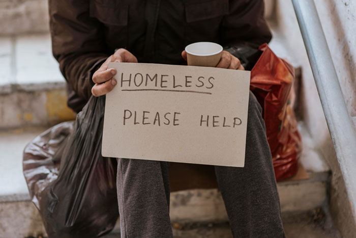 Person sitting on stairs, holding a Homeless Please Help sign and a cup, embodying the struggle of homelessness.