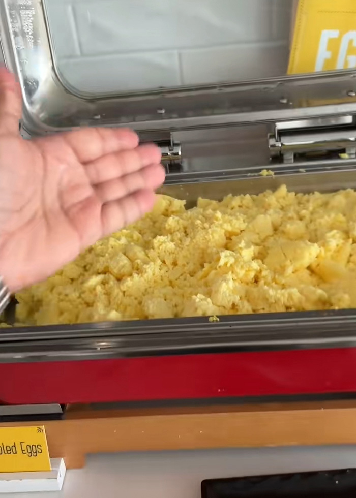 Uma mão paira sobre uma travessa cheia de ovos de bufê desagradáveis ​​e desajeitados, fazendo com que os hóspedes do hotel renunciem ao café da manhã grátis.