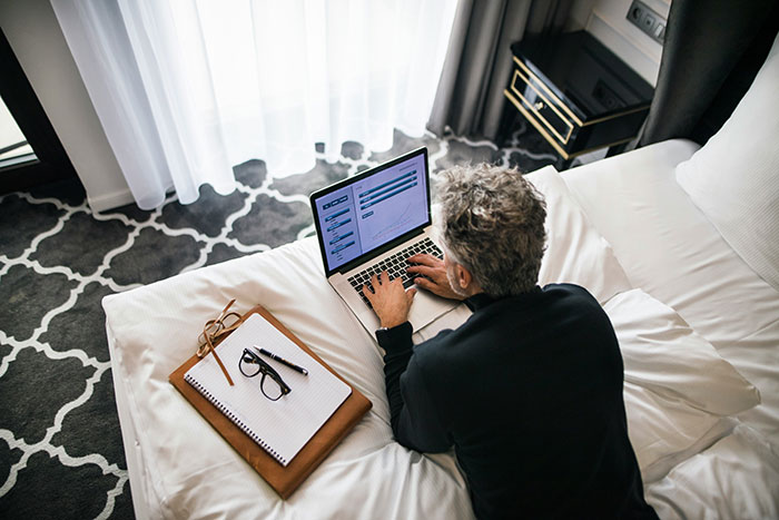 Person using laptop on hotel bed with notebook and glasses nearby, illustrating tips for an elevated and stress-free hotel stay.