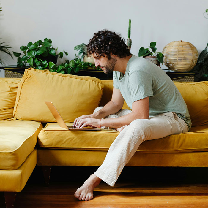 A man using a laptop on a yellow couch surrounded by plants, illustrating tips for an elevated and stress-free hotel stay.