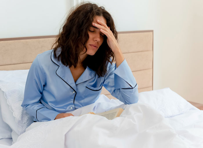 A woman in blue pajamas sits in bed, hand on her forehead, looking distressed. She is heartbroken after a cervical cancer diagnosis.