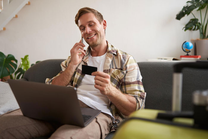 A smiling husband manages finances on his laptop, holding a credit card and talking on the phone.