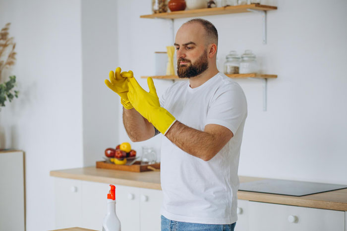 Bearded husband wearing yellow cleaning gloves in a kitchen, addressing house hoarder issues and finances.