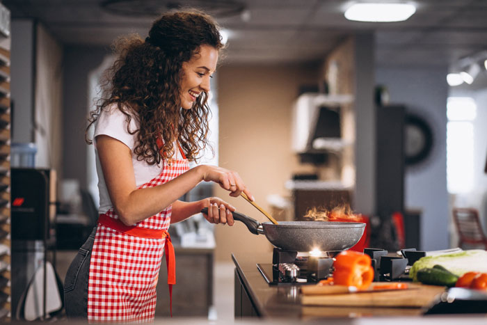 Woman cooking in kitchen wearing red apron, smiling while stirring food on stove, illustrating man outburst over mustard issue.