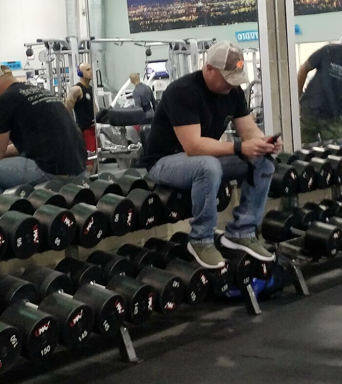 Man sitting on dumbbells at the gym, distracted by phone, showcasing unusual gym behavior and Darwin Award contender moments.