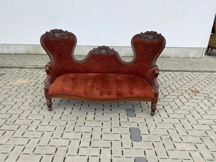 Antique red velvet sofa with ornate wooden carvings placed outdoors on a paved surface, elevator pit surroundings implied.