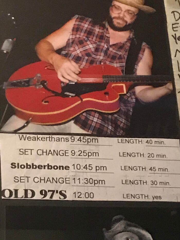 Man in a plaid sleeveless shirt playing red guitar at a live music event with a schedule showing band set times.