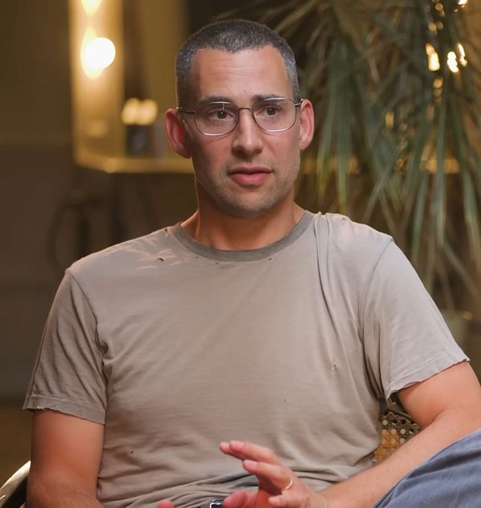 Man wearing glasses and a worn beige t-shirt speaking indoors with a plant in the background about main character syndrome.