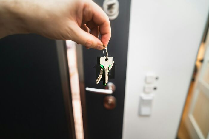 A hand holds house keys on a ring with a house-shaped tag. This image might relate to MIL stories.