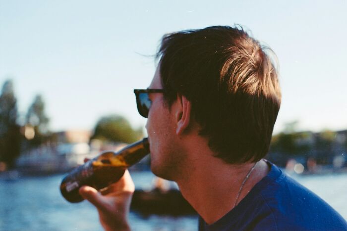 Man wearing sunglasses drinking from a bottle by the water, reflecting on normal family activities that seem bizarre now.