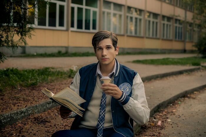 Young man displaying intelligence reading a book and drinking coffee outside near a school building in autumn.