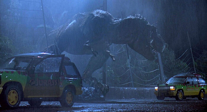 A massive T-Rex looming over a broken fence and two Jurassic Park vehicles in heavy rain, creating disturbing sounds.