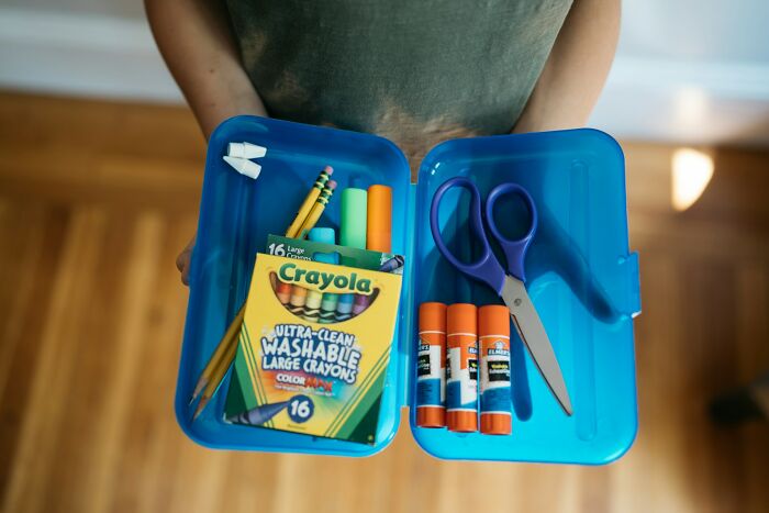 Person holding a blue container with crayons, glue sticks, scissors, and pencils, items TSA may confiscate from travelers