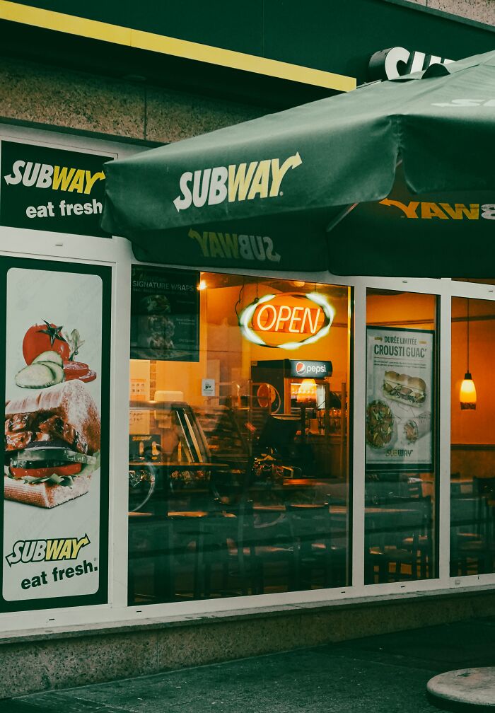 Subway restaurant storefront with open sign, green awning, and visible seating inside during evening hours.