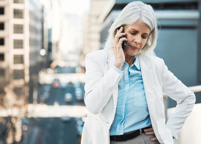 Middle-aged woman in white jacket talking on phone outdoors looking concerned about kid work mother fired issue. Middle-aged woman in white jacket talking on phone outdoors looking concerned about kid work mother fired issue.