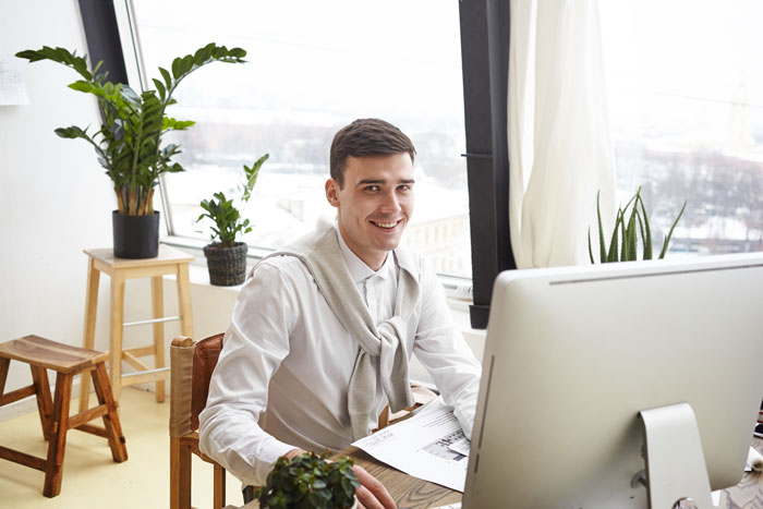 Young man working from home on computer, smiling, with plants in bright room, illustrating kid work mother fired context. Young man working from home on computer, smiling, with plants in bright room, illustrating kid work mother fired context.